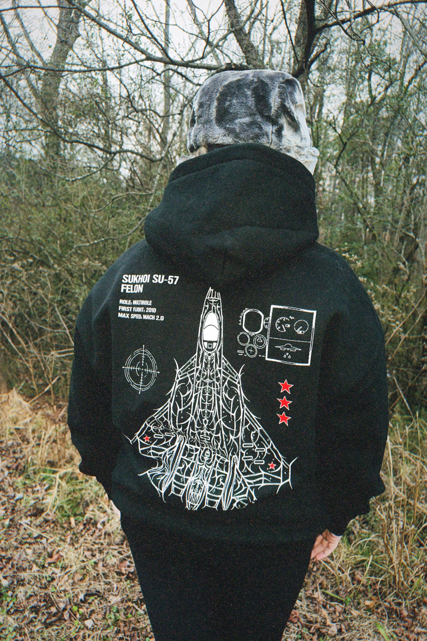 Person outdoors wearing black hoodie with aviation-inspired Sukhoi SU-57 jet graphic and text on back