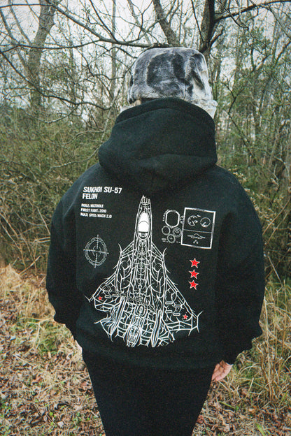 Person outdoors wearing black hoodie with aviation-inspired Sukhoi SU-57 jet graphic and text on back