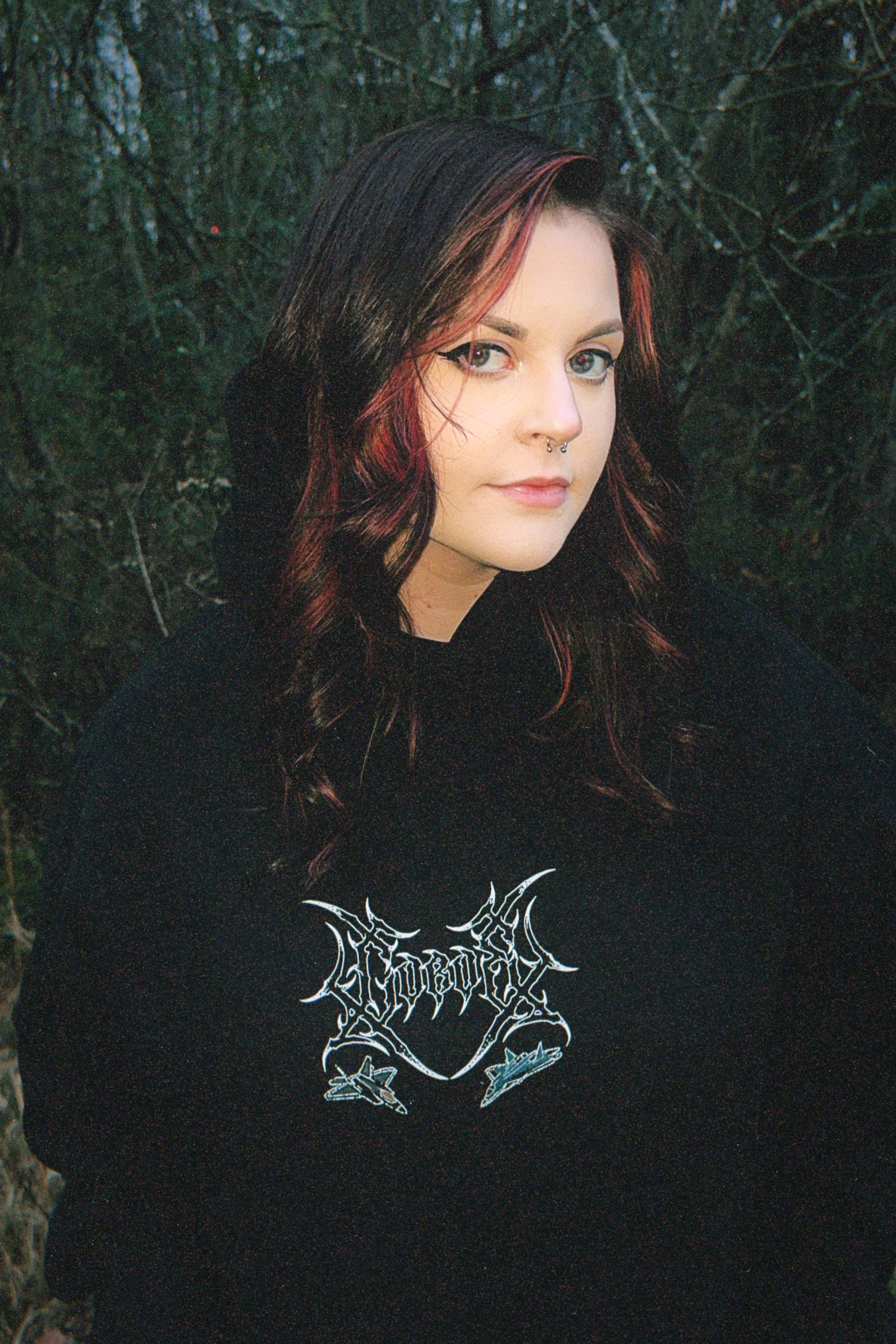 Woman in black graphic hoodie with aviation-inspired logo, outdoors in forest setting