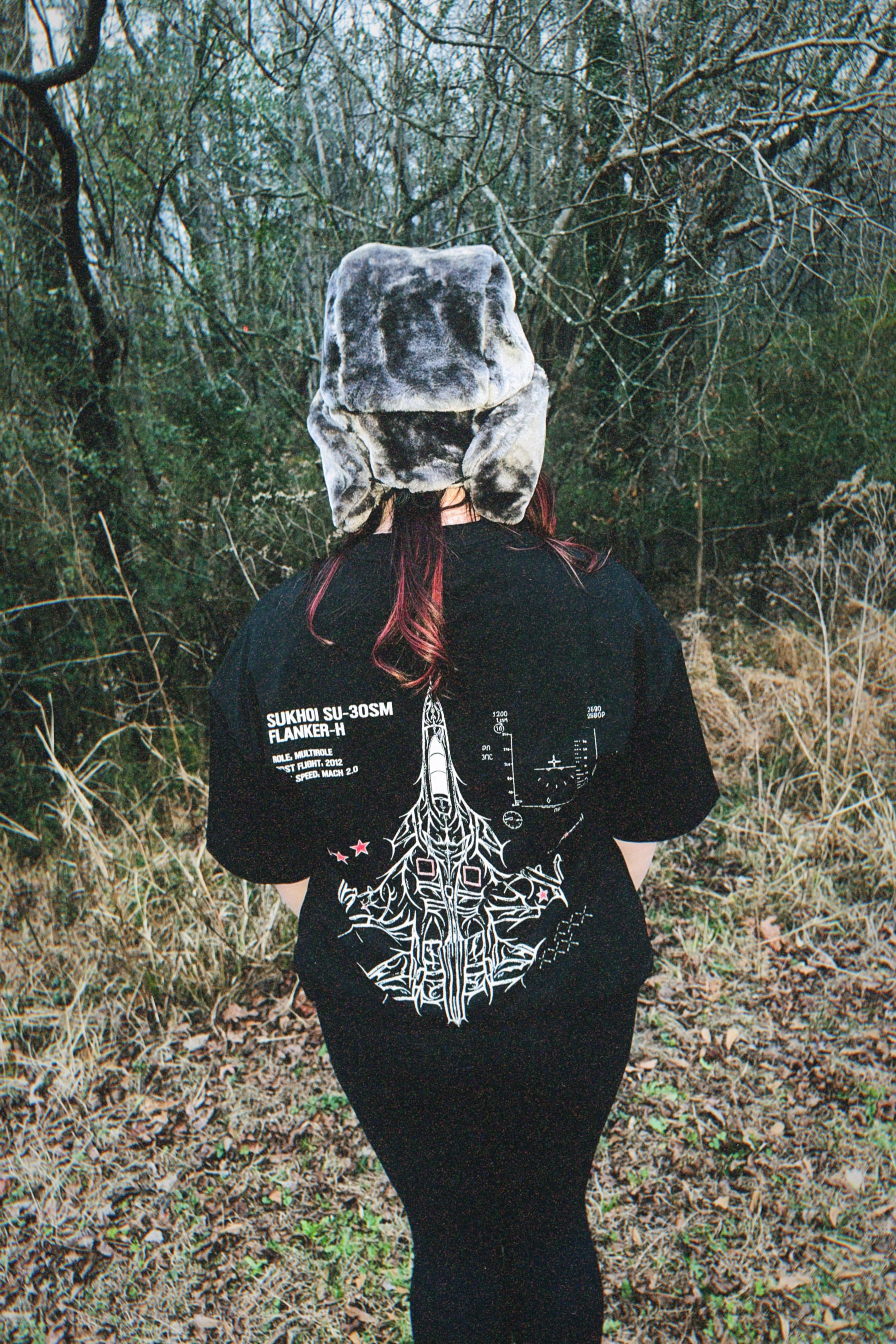 Person wearing aviation-inspired streetwear t-shirt and fur hat in wooded outdoor setting