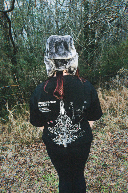 Person wearing aviation-inspired streetwear t-shirt and fur hat in wooded outdoor setting
