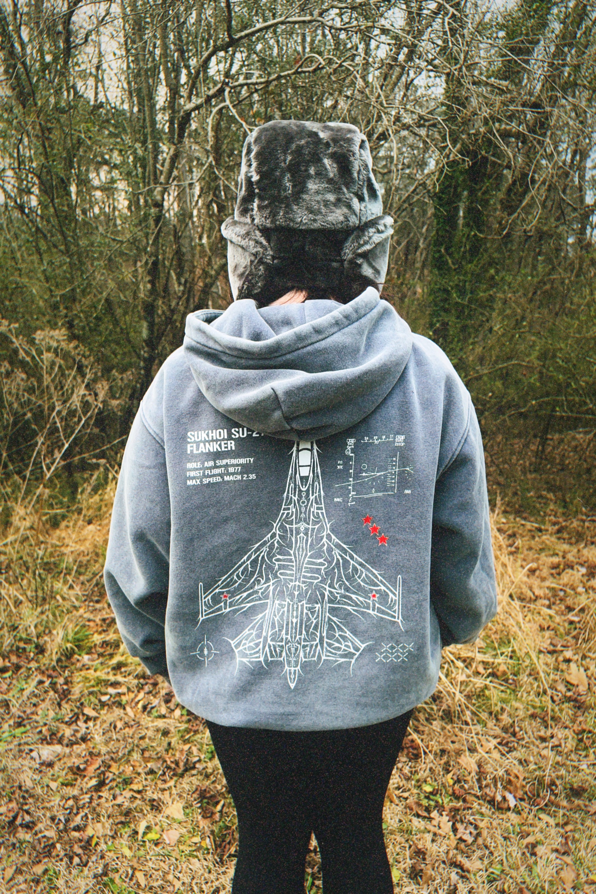 Person wearing a grey hoodie with aviation-inspired Sukhoi Su-27 jet line art in a forest