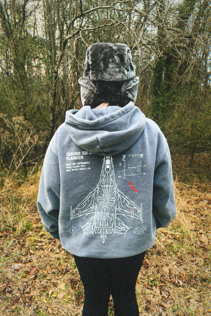 Person wearing a grey hoodie with aviation-inspired Sukhoi Su-27 jet line art in a forest