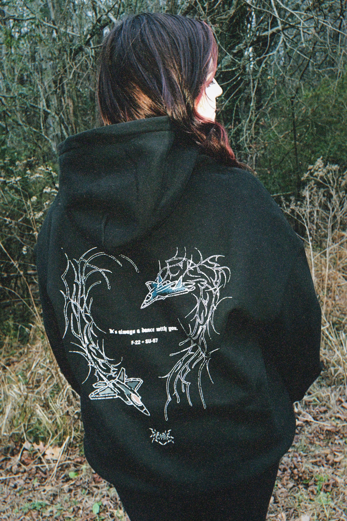 Person in black hoodie with aviation fighter jet and thorn heart graphic, outdoor setting