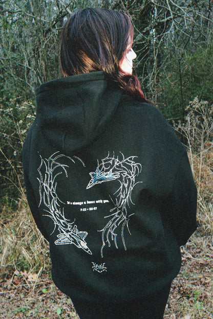 Person in black hoodie with aviation fighter jet and thorn heart graphic, outdoor setting