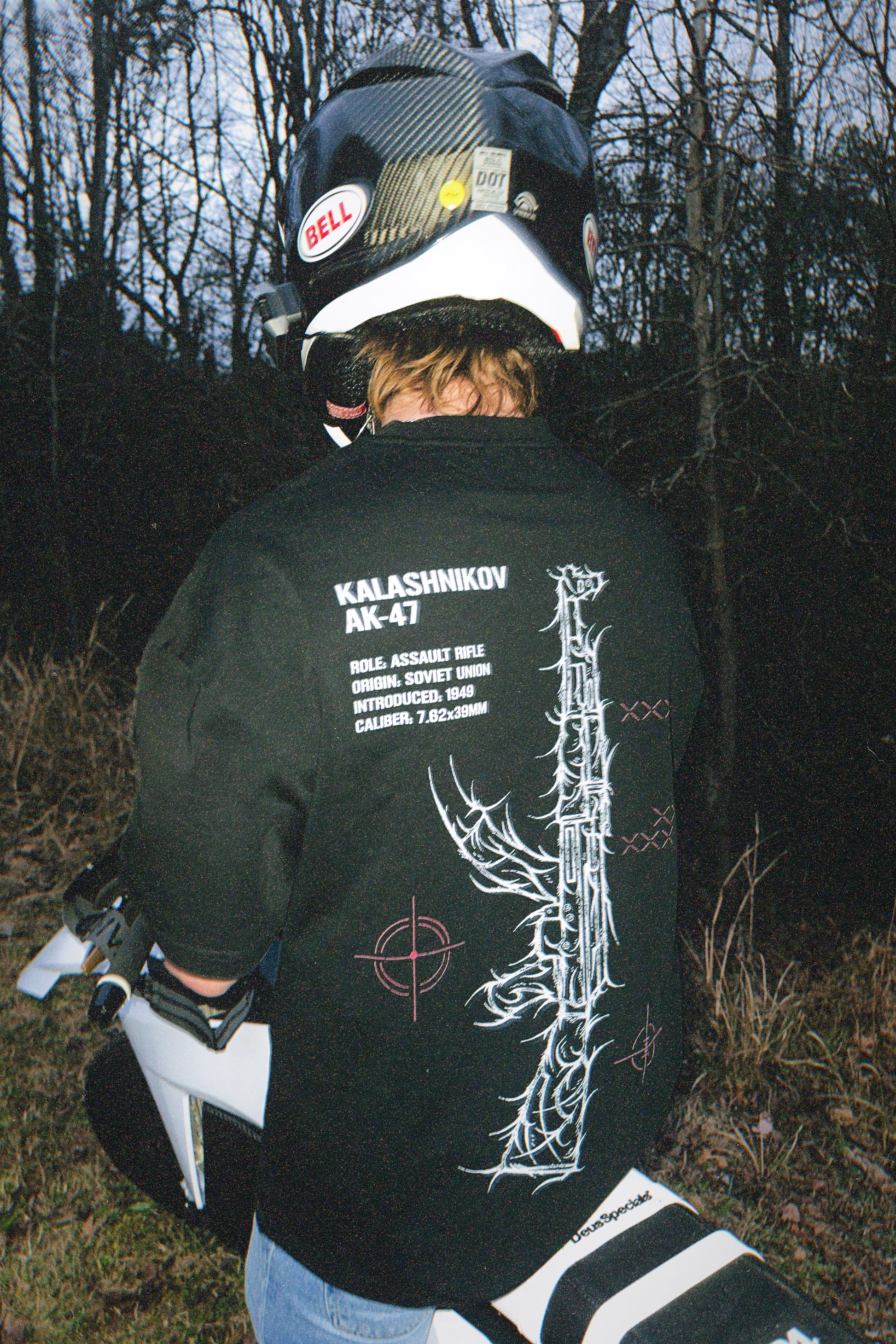 Person wearing black tactical tee with graphic rifle design, sitting on a motorcycle outdoors