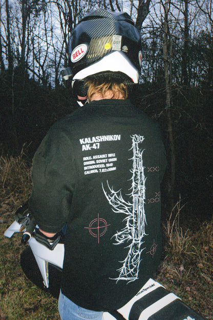 Person wearing black tactical tee with graphic rifle design, sitting on a motorcycle outdoors