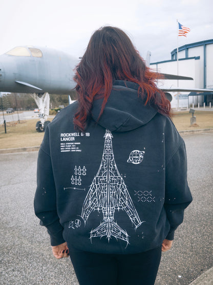 Person wearing a black hoodie with Rockwell B-1B Lancer aircraft blueprint graphic, standing in front of a military jet outdoors.