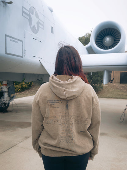 Person in tan aviation hoodie with aircraft blueprint stands near military jet outdoors