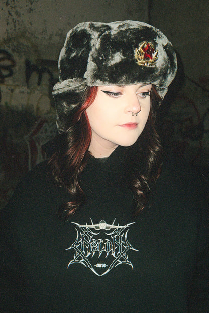 Person in a dark streetwear hoodie with aviation-inspired graphic and furry hat, urban background