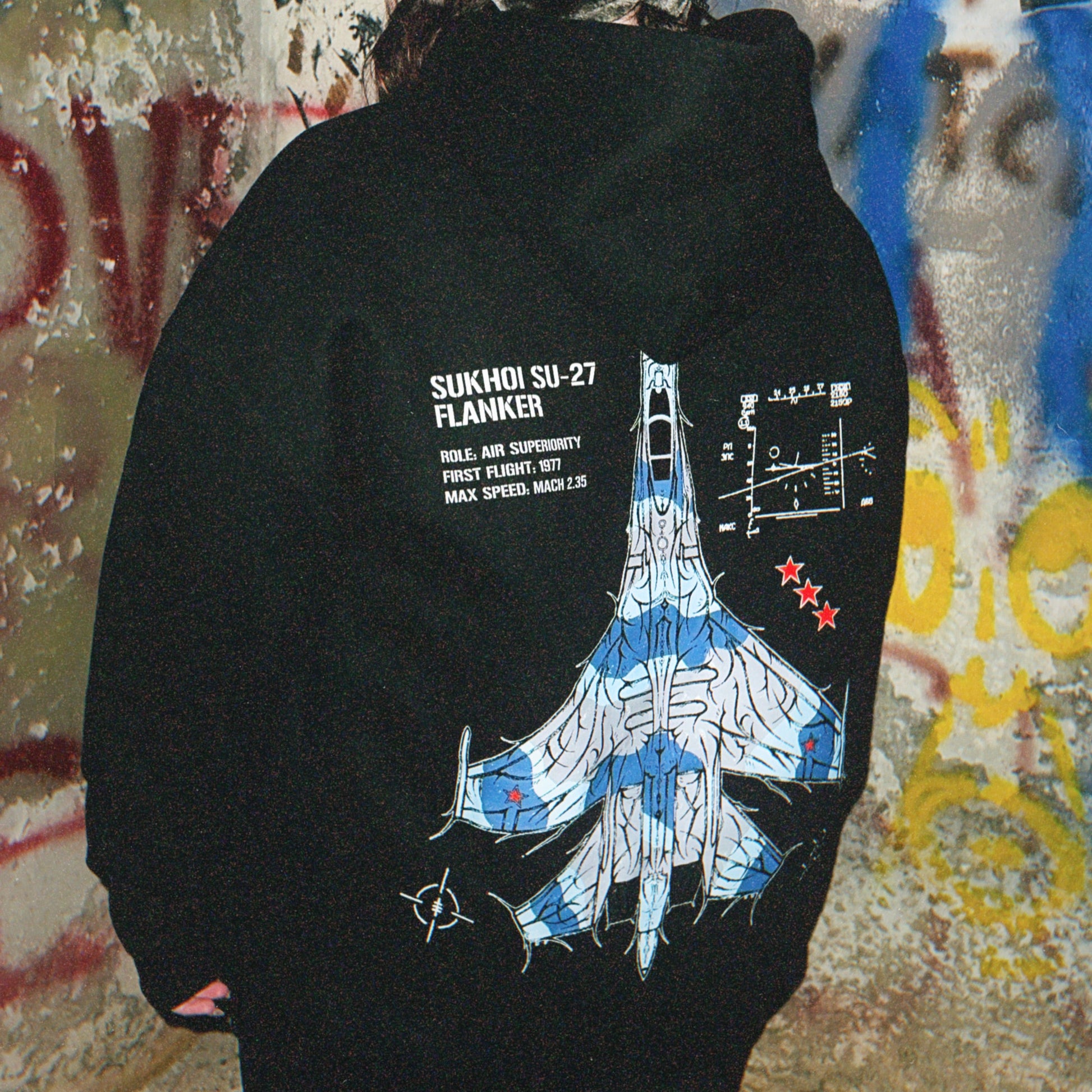 Back of a black hoodie with Sukhoi SU-27 fighter jet graphic and specs, worn in urban graffiti setting.