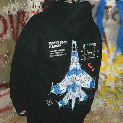 Back of a black hoodie with Sukhoi SU-27 fighter jet graphic and specs, worn in urban graffiti setting.