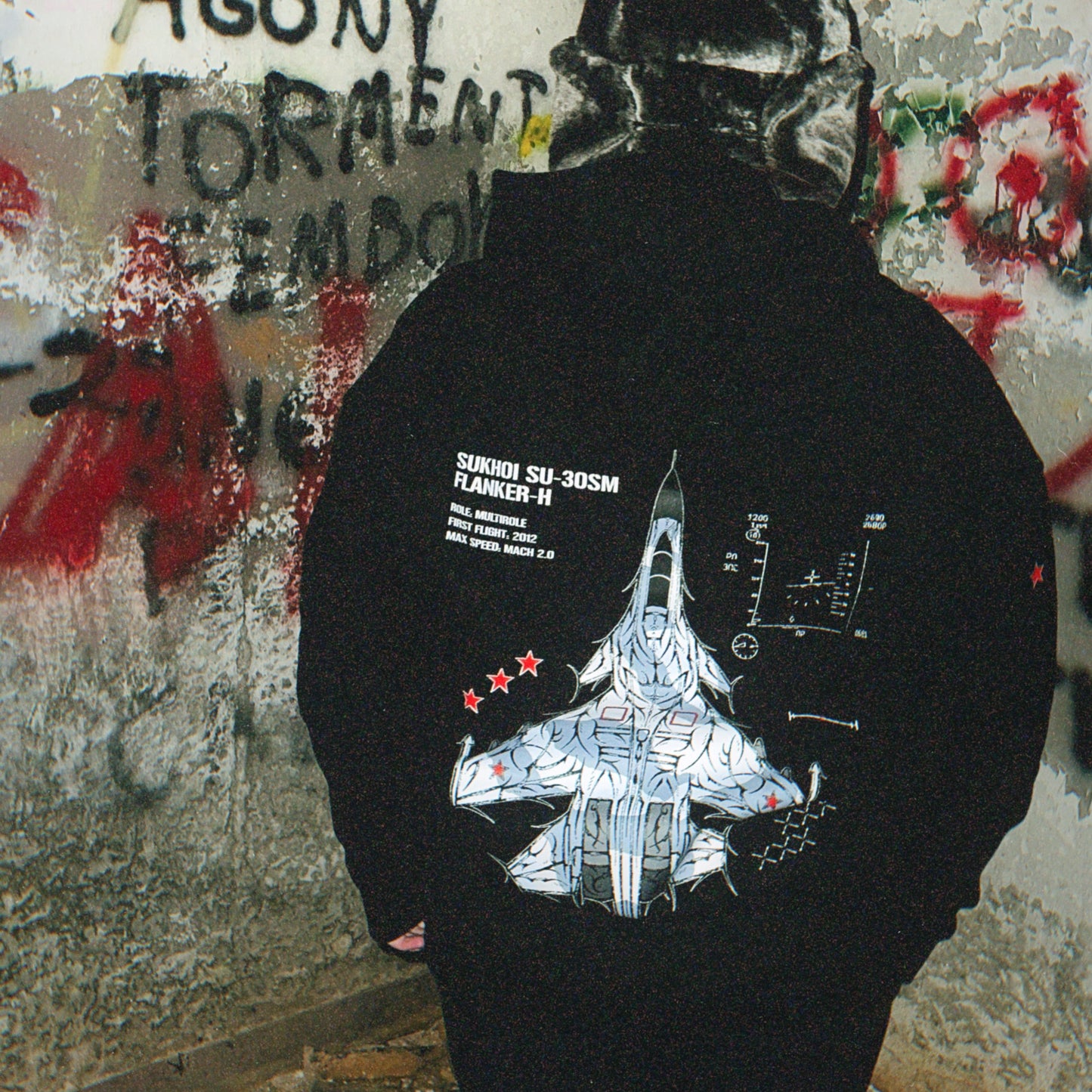 Back of a black hoodie featuring a Sukhoi SU-30SM fighter jet graphic and aviation details, worn outdoors against graffiti wall