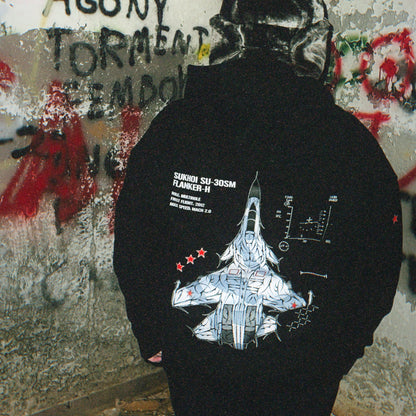 Back of a black hoodie featuring a Sukhoi SU-30SM fighter jet graphic and aviation details, worn outdoors against graffiti wall