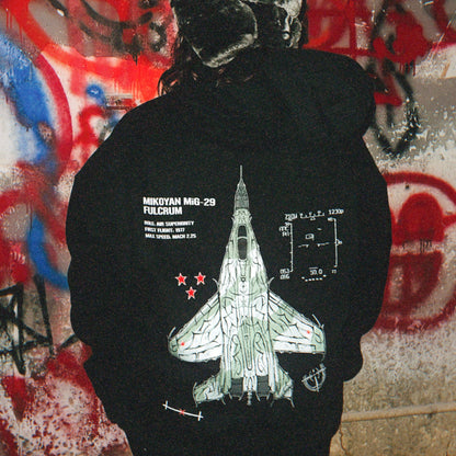 Person wearing aviation-inspired hoodie with MiG-29 jet graphic and technical details against graffiti wall