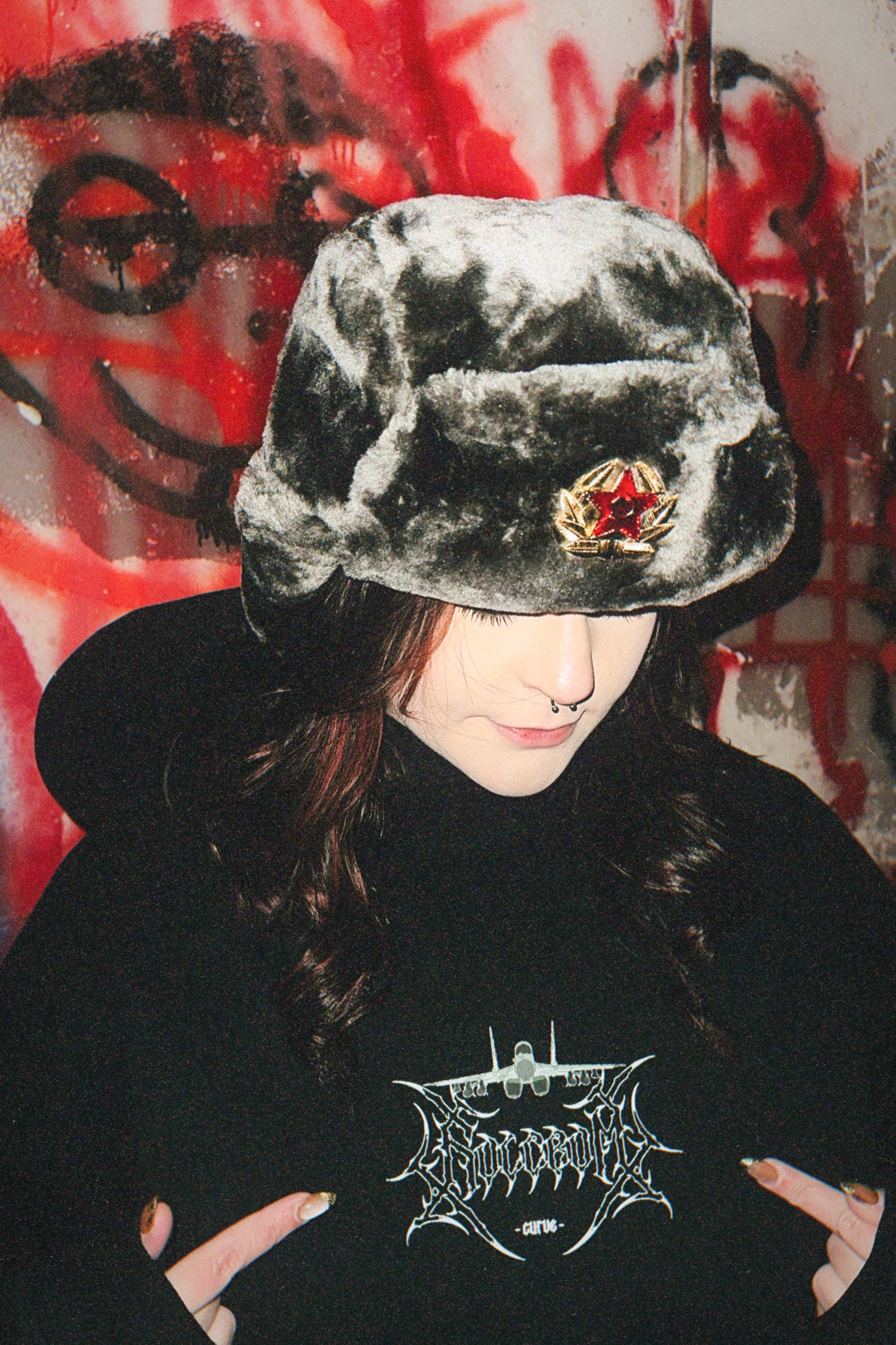 Person wearing gray faux fur ushanka and black hoodie with aviation graphic, graffiti background