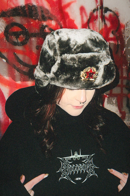 Person wearing gray faux fur ushanka and black hoodie with aviation graphic, graffiti background
