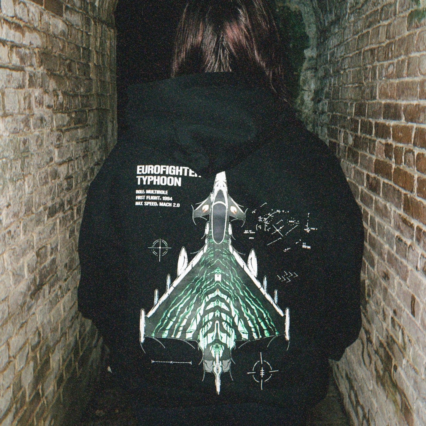 Person in alley wears black hoodie with Eurofighter Typhoon jet graphic, aviation streetwear
