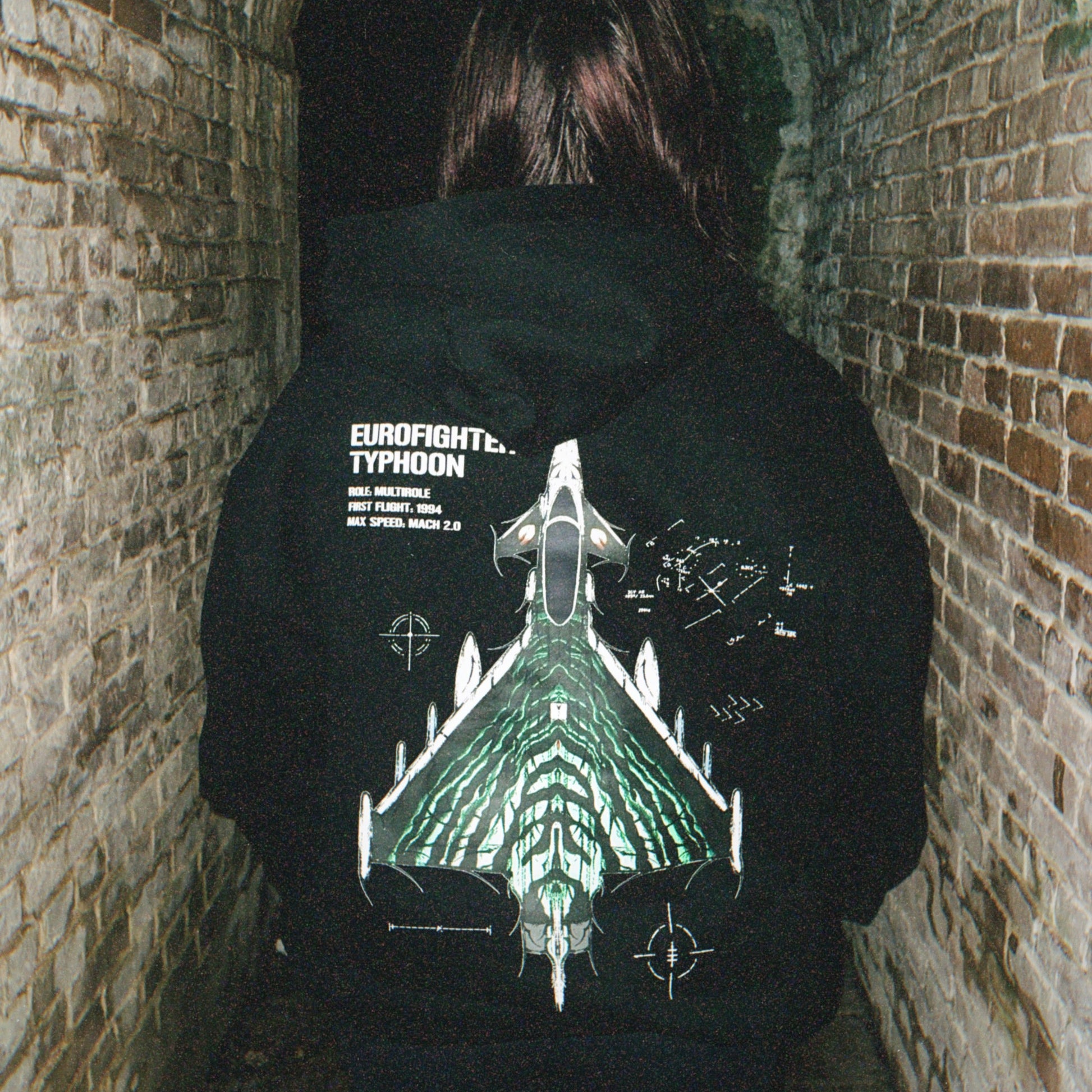 Person in alley wears black hoodie with Eurofighter Typhoon jet graphic, aviation streetwear