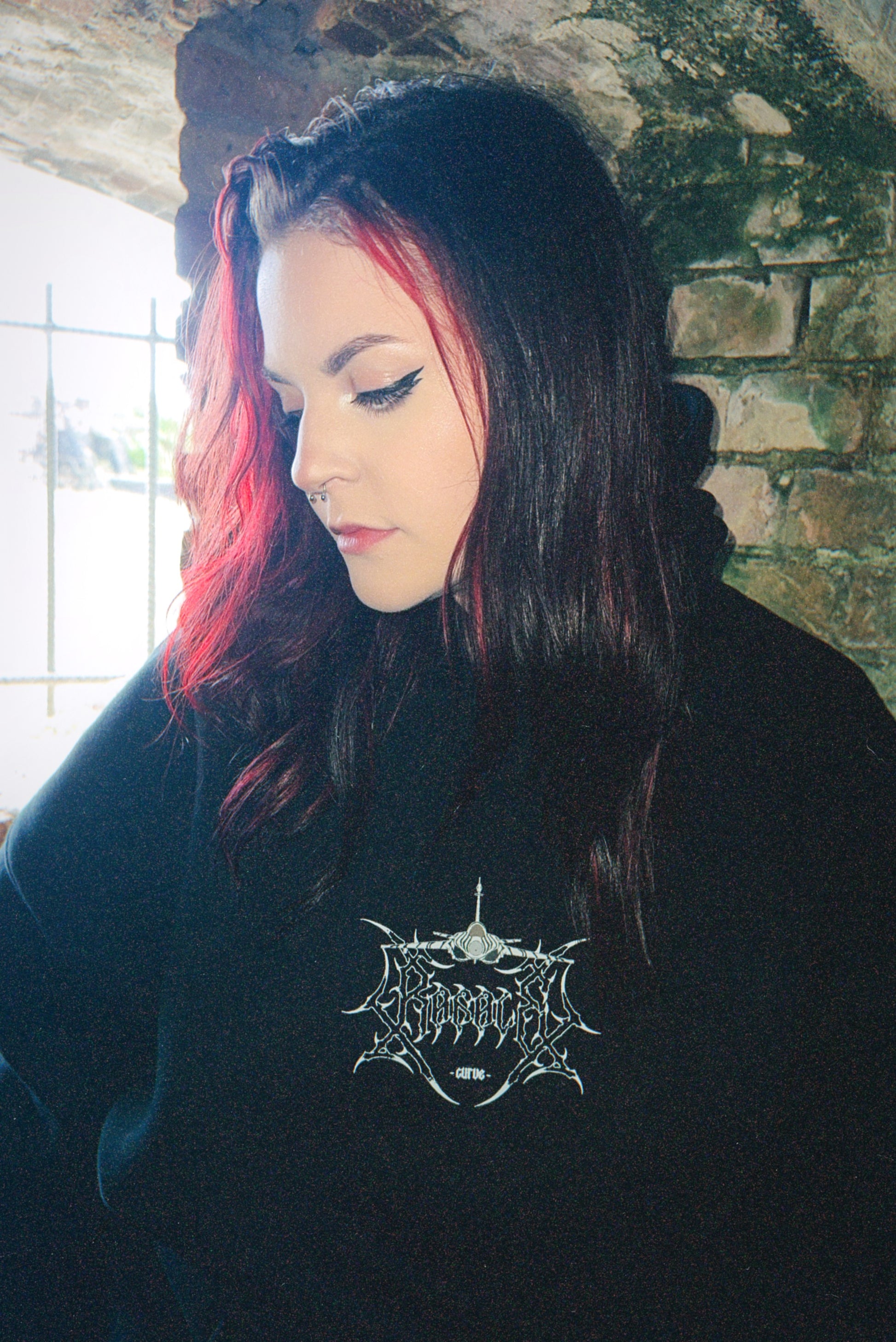 Woman with dark hair and red highlights wearing black streetwear hoodie with graphic logo, standing against a brick wall.