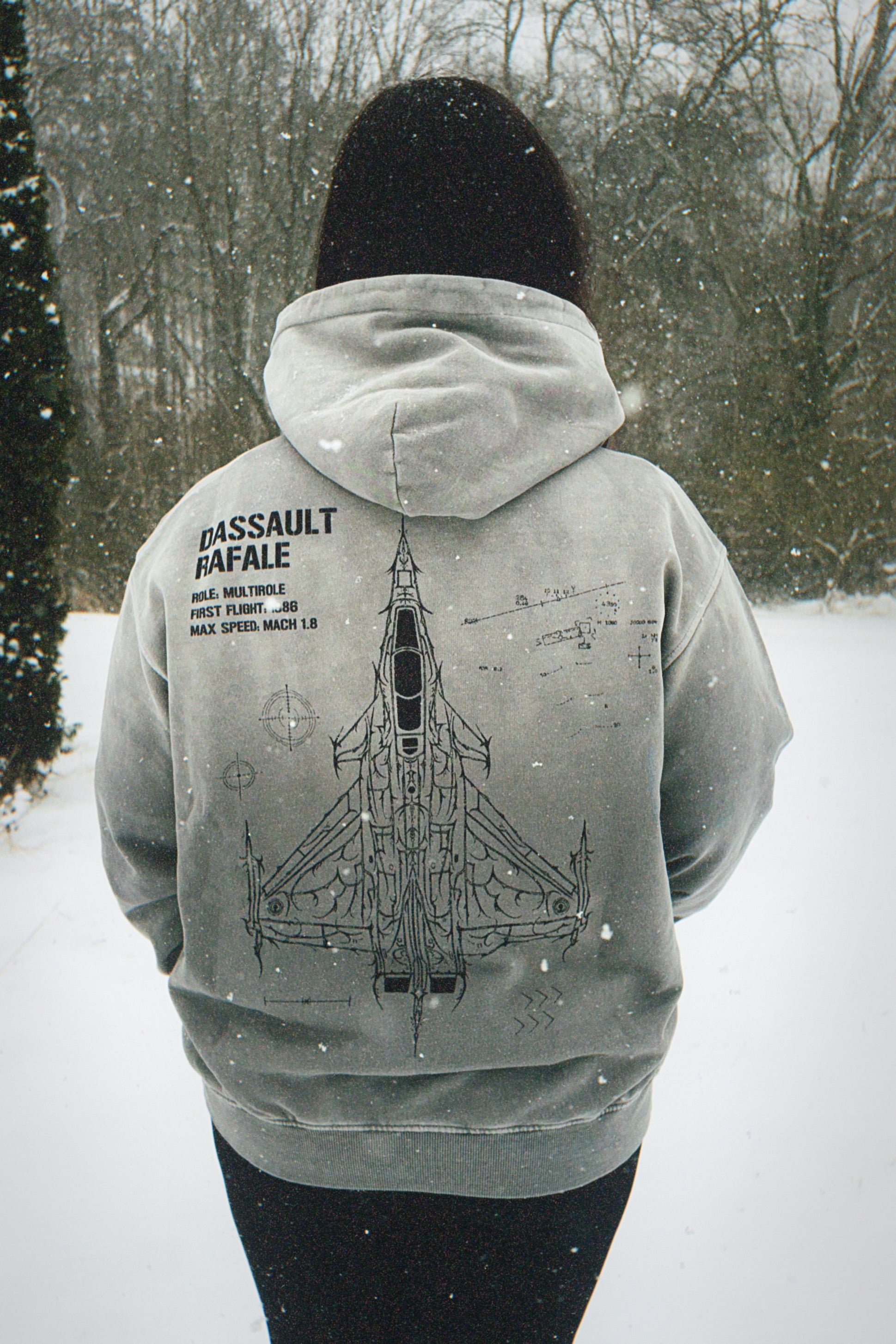Person wearing a grey hoodie with Dassault Rafale jet graphic and specs, outdoors in snow