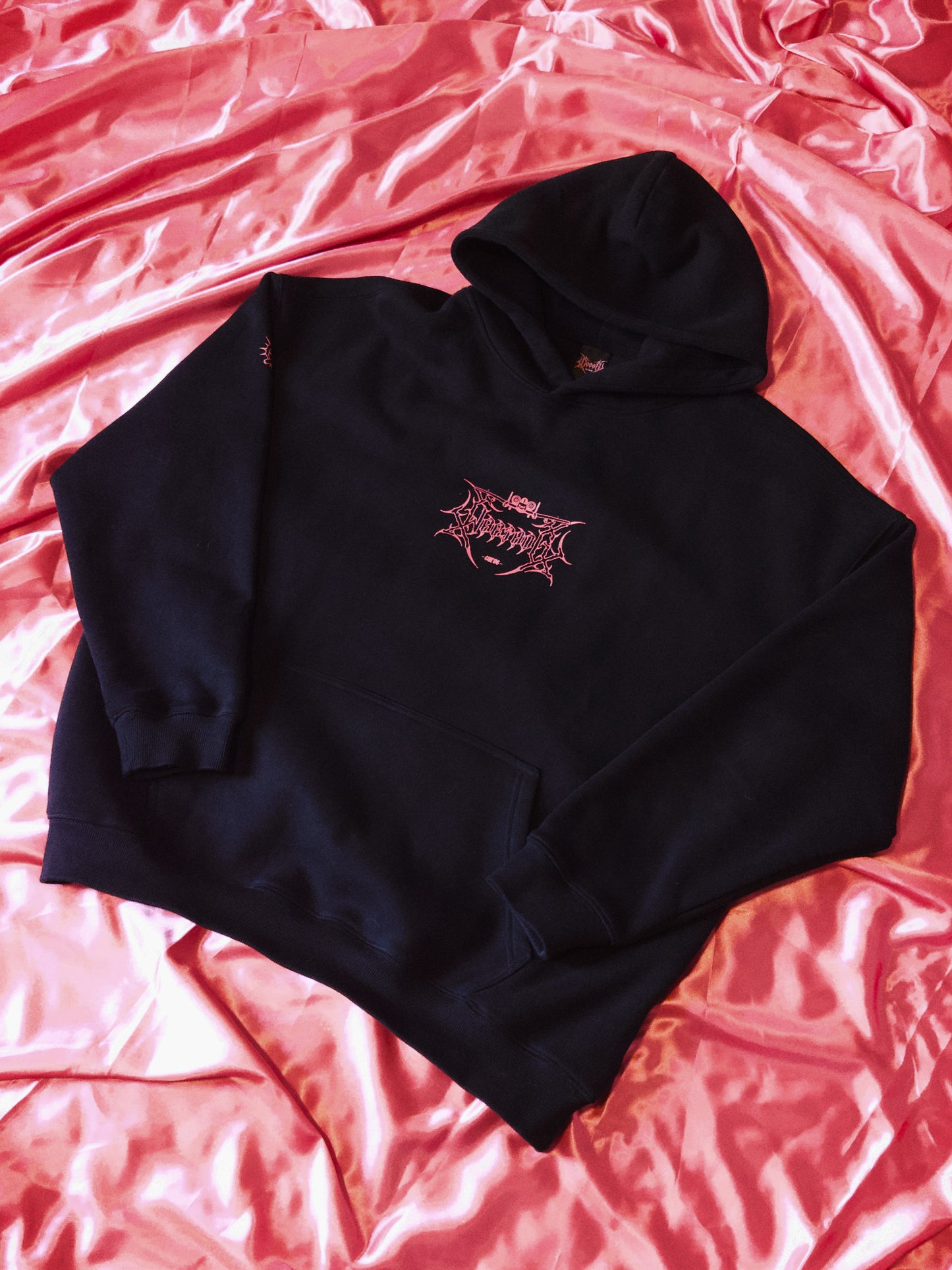 Black streetwear hoodie with pink graphic design on pink satin fabric background.