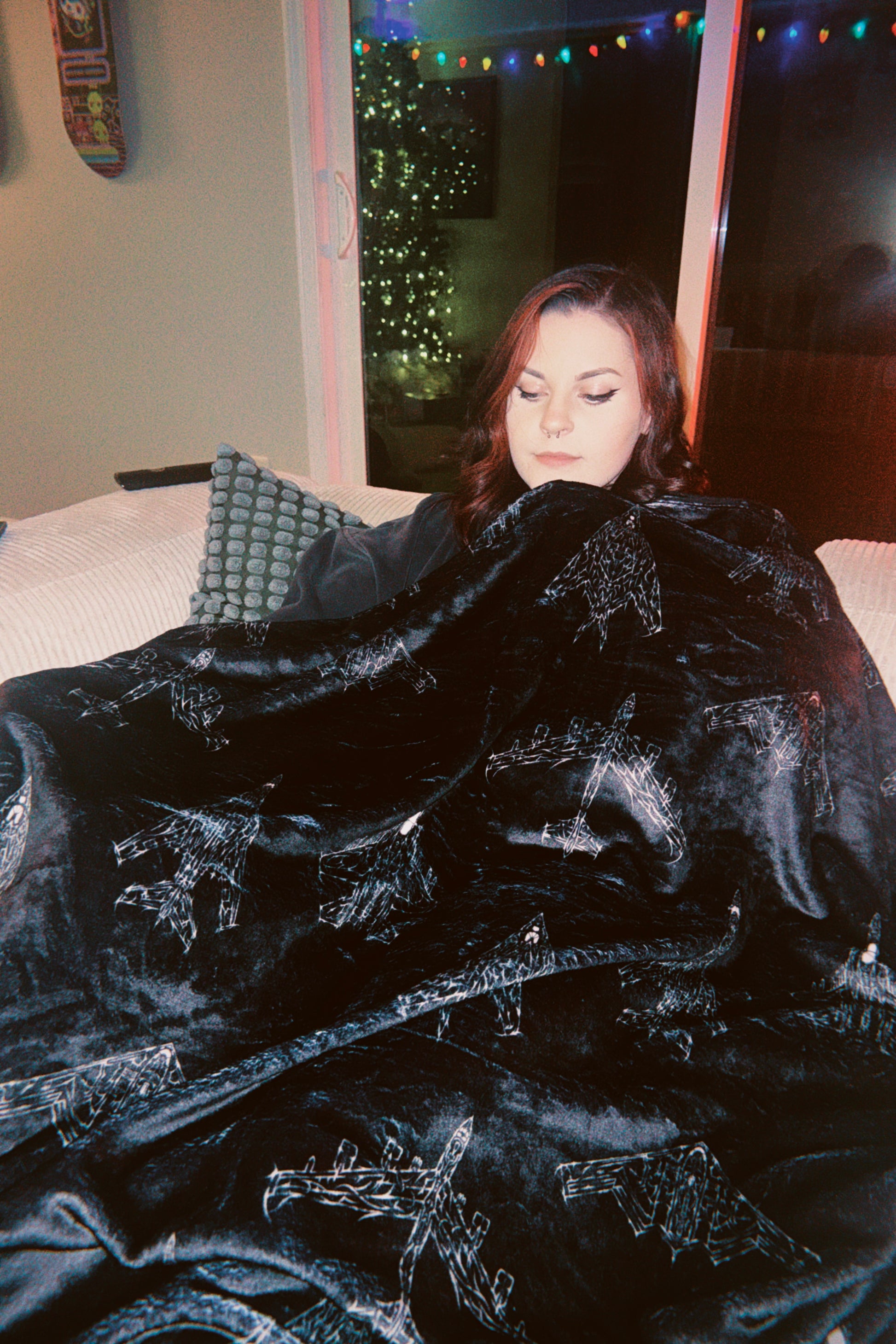 Woman on couch with black aviation-themed streetwear blanket, holiday lights in background