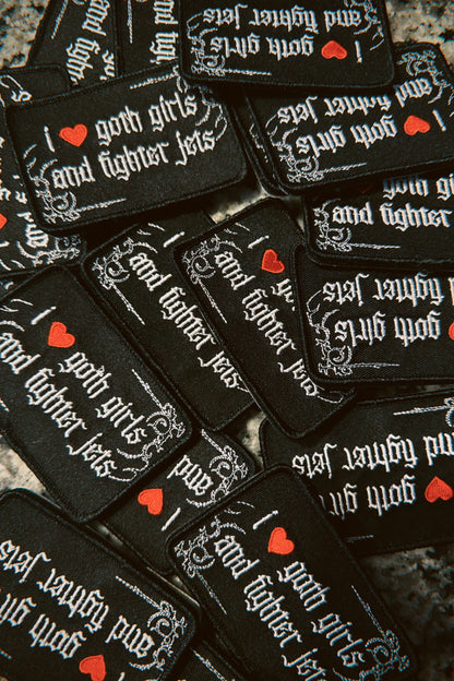 Pile of black embroidered patches with gothic text reading 'I love goth girls and fighter jets'.